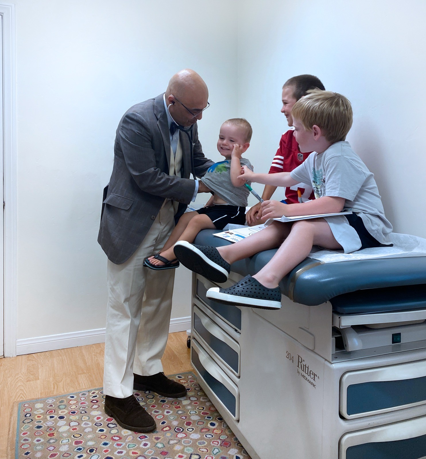 Dr. Mehr listening with a stethoscope while three children sit together on an examination table.